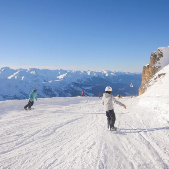 A beginner snowboarder riding a wide, sunlit piste above the valley.