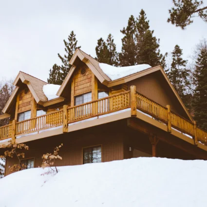 A timber alpine chalet with deep snow on its roof, framed by pine trees.