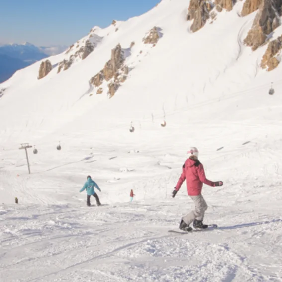 Two snowboarders linking turns down a groomed run beneath rocky peaks.